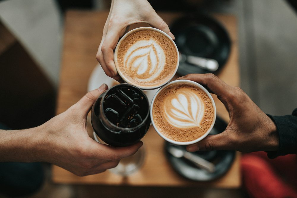 Three cups of coffee held by three hands over a wooden table — two flat whites with latte art, and a cold brew.